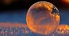 Close-up shot of a frozen bubble with warm reflections resting on a snowy surface at twilight.